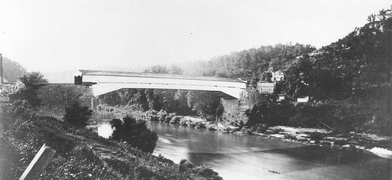 A wooden bridge spans a river, with cliffs covered with trees on both shores of the river during the Civil War.