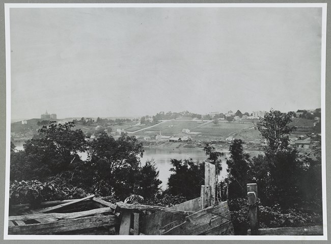 View across water source with buildings, trees, and fields in the background during the Civil War.