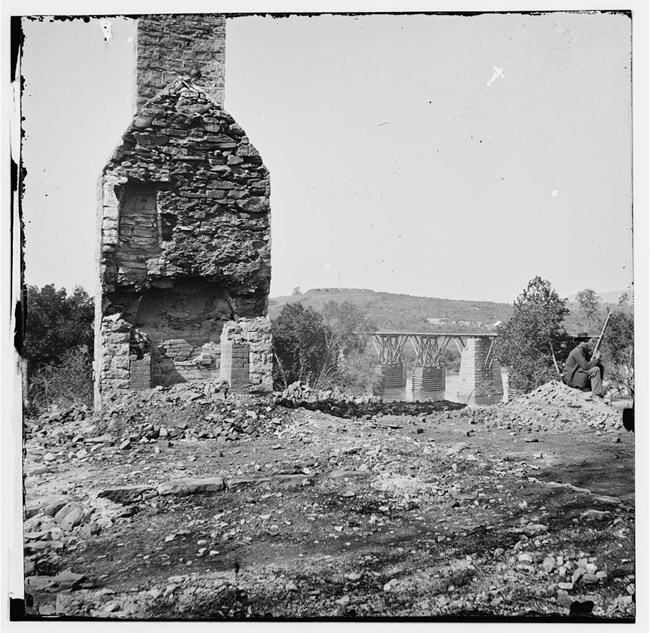 Brick chimney in the foreground and bridge over water source with a fort in the background during the Civil War.