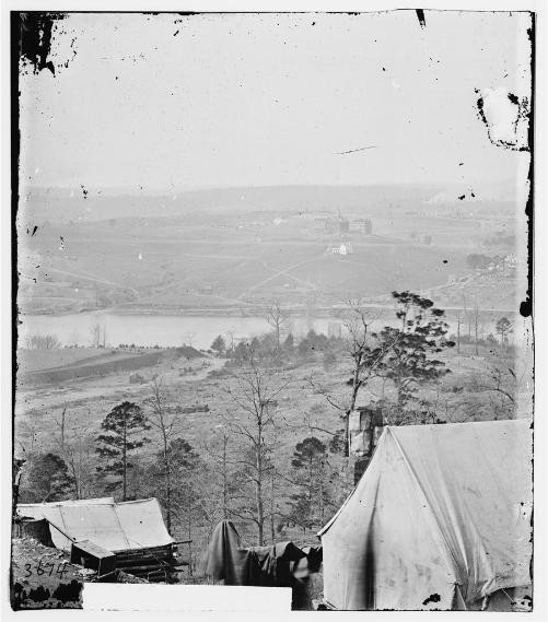 Open ground and building across a water source with tents in the foreground during the Civil War.