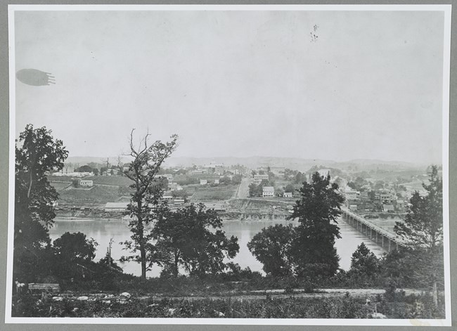 Tall trees in the foreground with a bridge crossing water during the Civil War. Buildings and roads can be seen in the background.