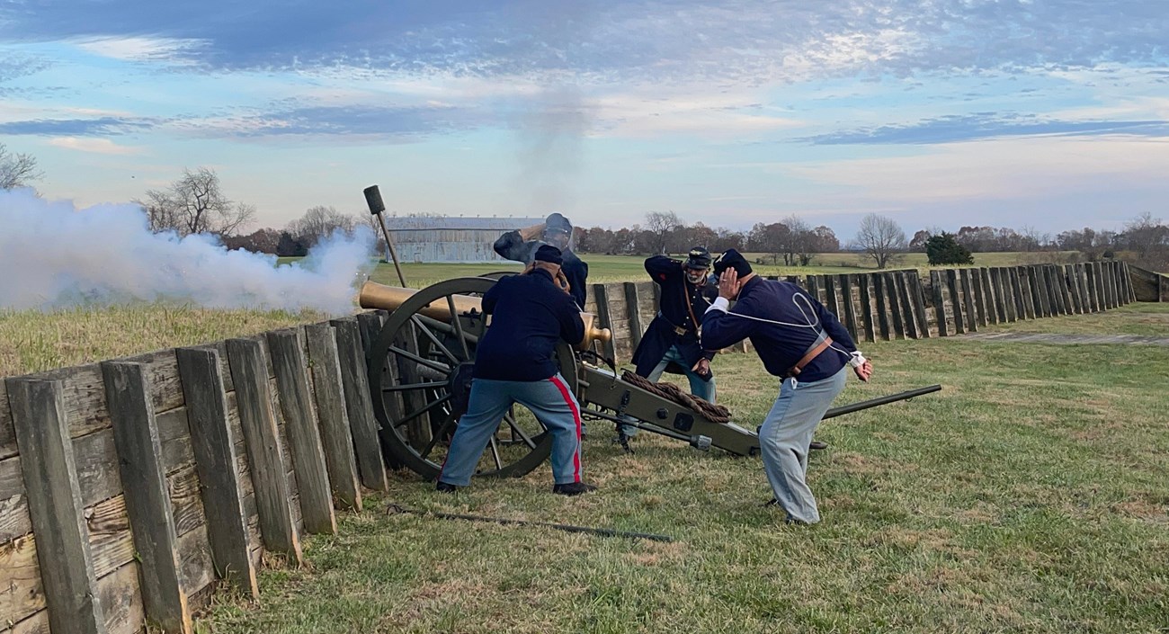 12th United States Colored Heavy Artillery, in blue uniforms, firing a cannon at Fort Putnam. Smoke is coming out of the cannon. The fort is covered with grass and wood.