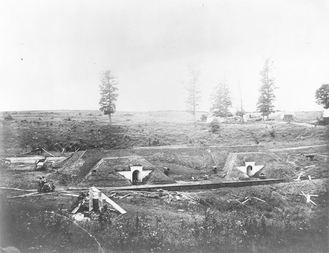 Large mound of earth with two entrances in the foreground and open countryside with a few trees in the background during the Civil War.