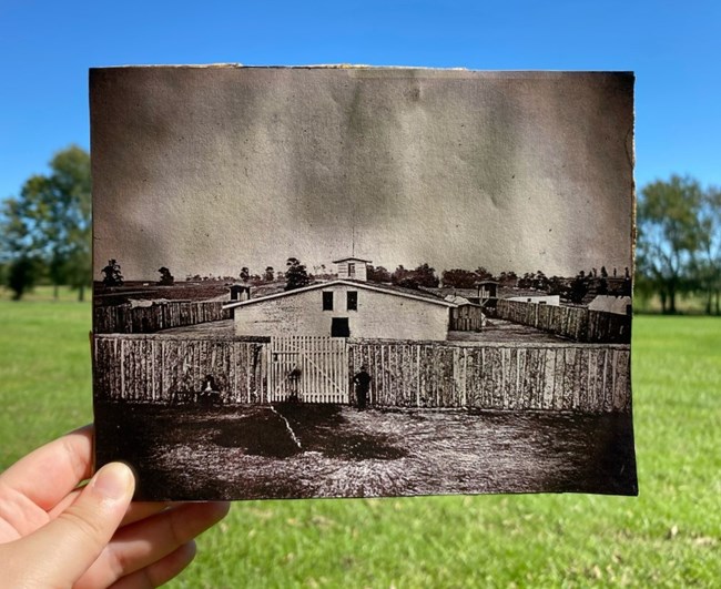 A hand is holding a historic image of a prison constructed of wood. Behind the image is green grass and trees, and a blue sky.