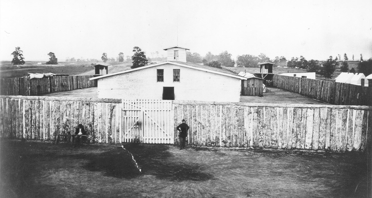 A wooden structure with a main building behind the fence with wooden towers during the Civil War. Trees be seen in the background.