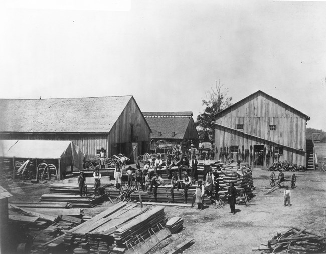 Large buildings in the background with over a dozen men posing while sitting and standing on a large stack of lumber during the Civil War.