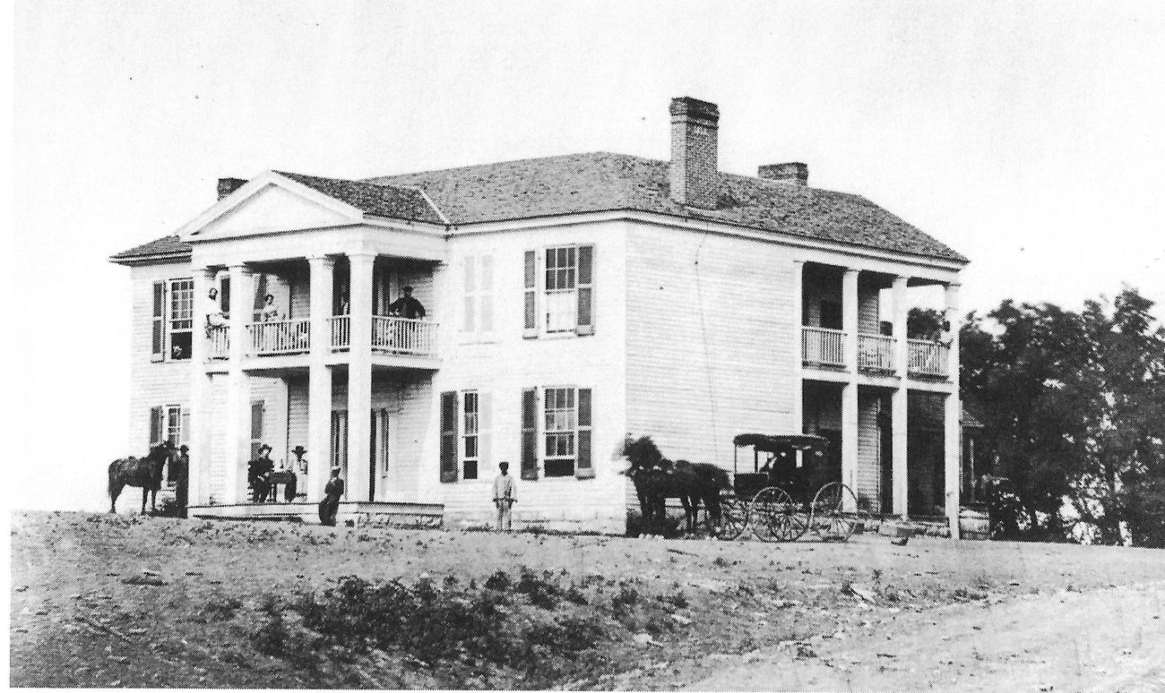 A white house with people standing and sitting on the front steps and second floor balcony during the Civil War. There are horses and a carriage next to the house. Trees can been seen on the right and a dirt road in the foreground.