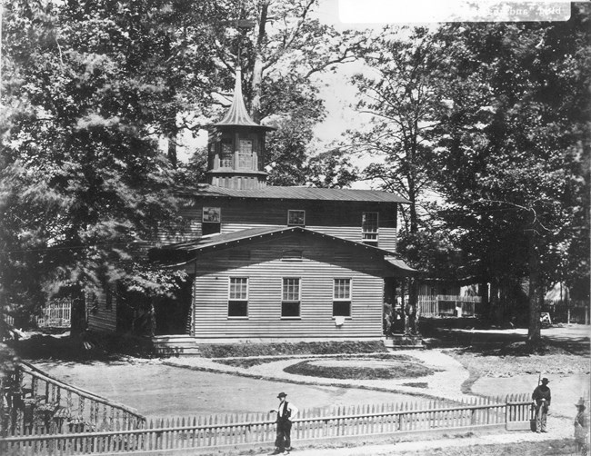 Two-story wooden building surrounded by trees and a fence in front.