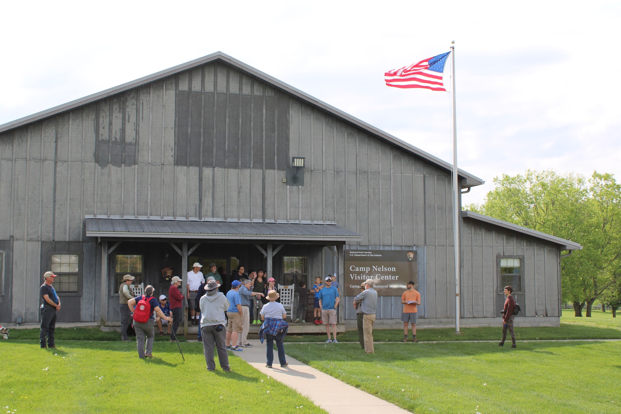 279858488_522287545992225_3576361351098737887_n Large grey building with several dozen people standing in front of it on patio and green grass.