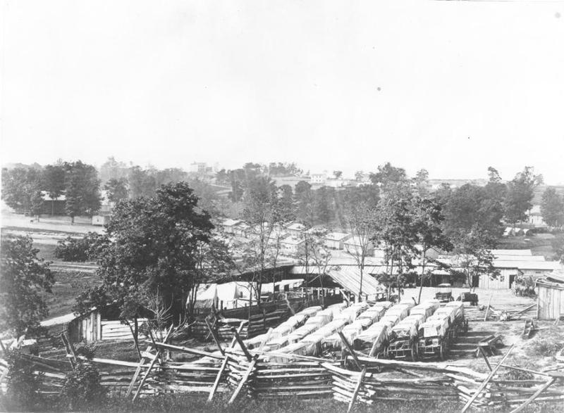 Large group of wagons surrounded by buildings, fences, and trees during the Civil War.
