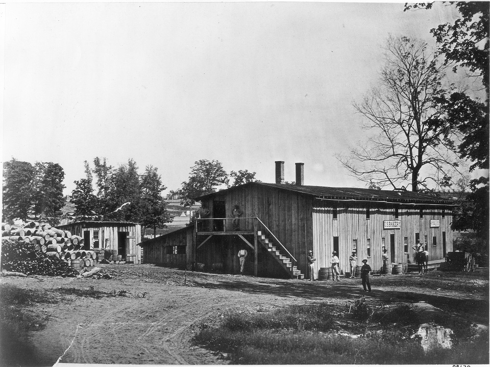 Civil War photograph of a large two story building that was the Bakery for Camp Nelson. There are 10 men and mounds of barrels outside the building.