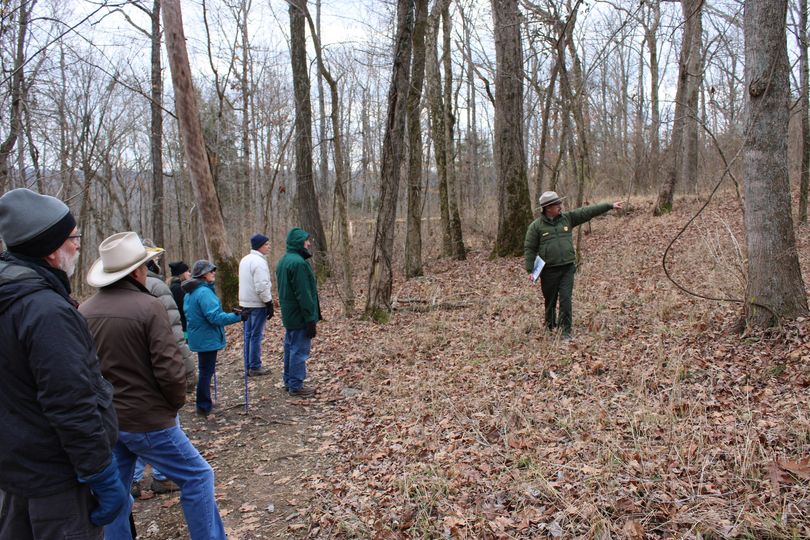 A park ranger pointing to a historic feature to a group of hikers in a forest area with an overlook in the background.