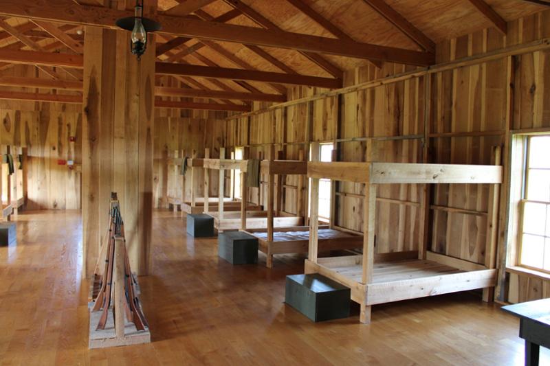 353422233_5d660015190390588_298106907652552090_n Interior of reconstructed wooden barracks building with double bunks and metal stove.