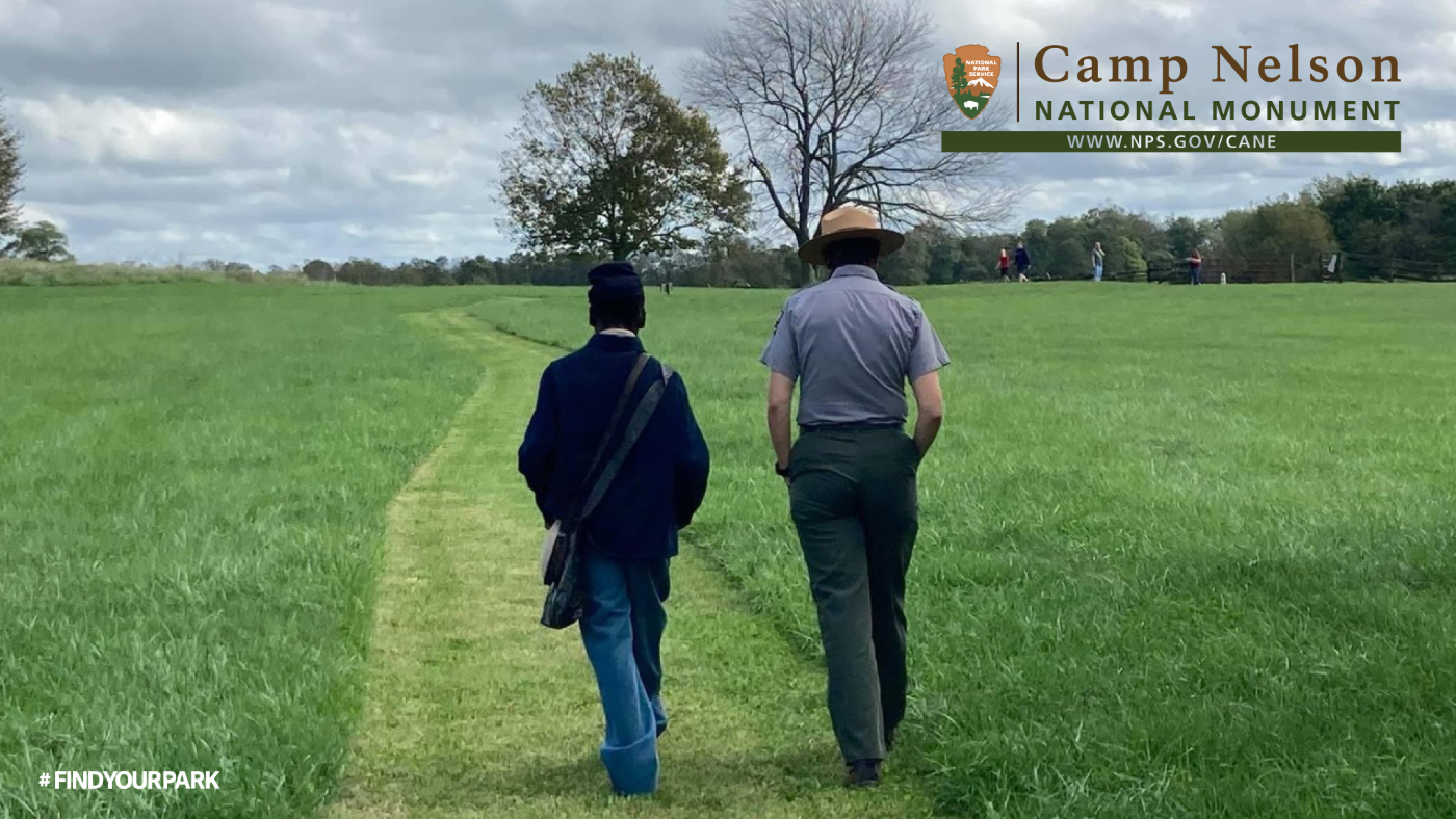 Ranger walking with visitor along grass trail with trees in the background.