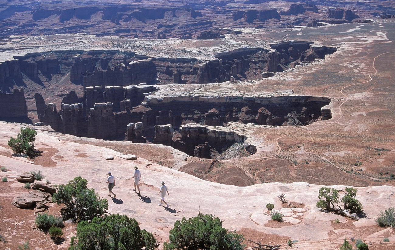 Hikers walk on a flat slickrock ledge on the rim of a huge canyon.