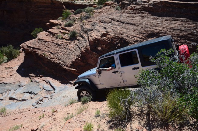 A white jeep drives down a narrow and steep slickrock road.