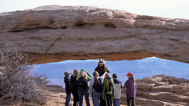 A park ranger talks to a group of students in front of a large sandstone arch. Canyons are visible through the arch beyond the group.