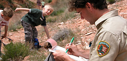 a man wearing a brown volunteer uniform writes on a notepad. A boy stands in the background a man wearing a brown volunteer uniform writes on a notepad. A boy stands in the background