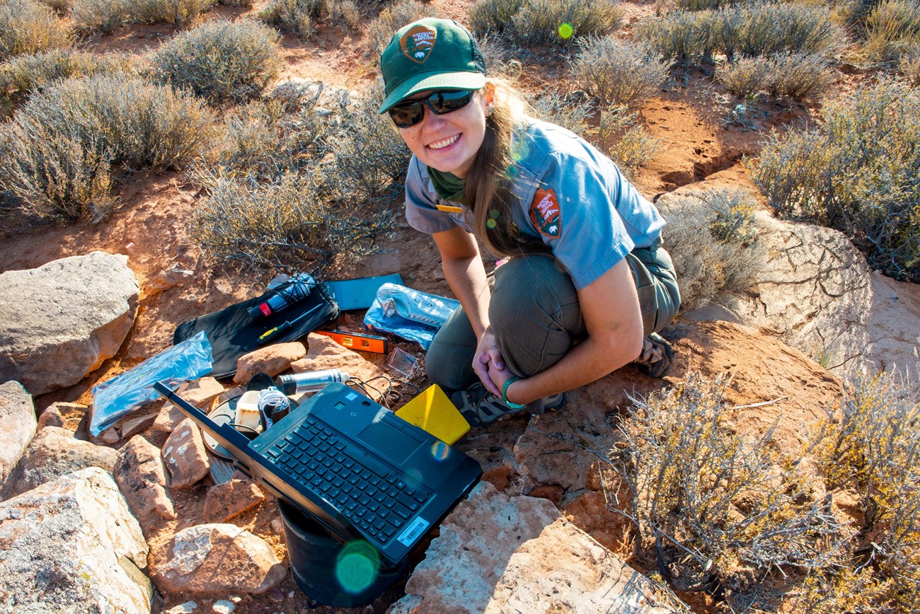 A researcher with various tools and a laptop collects data in the field.