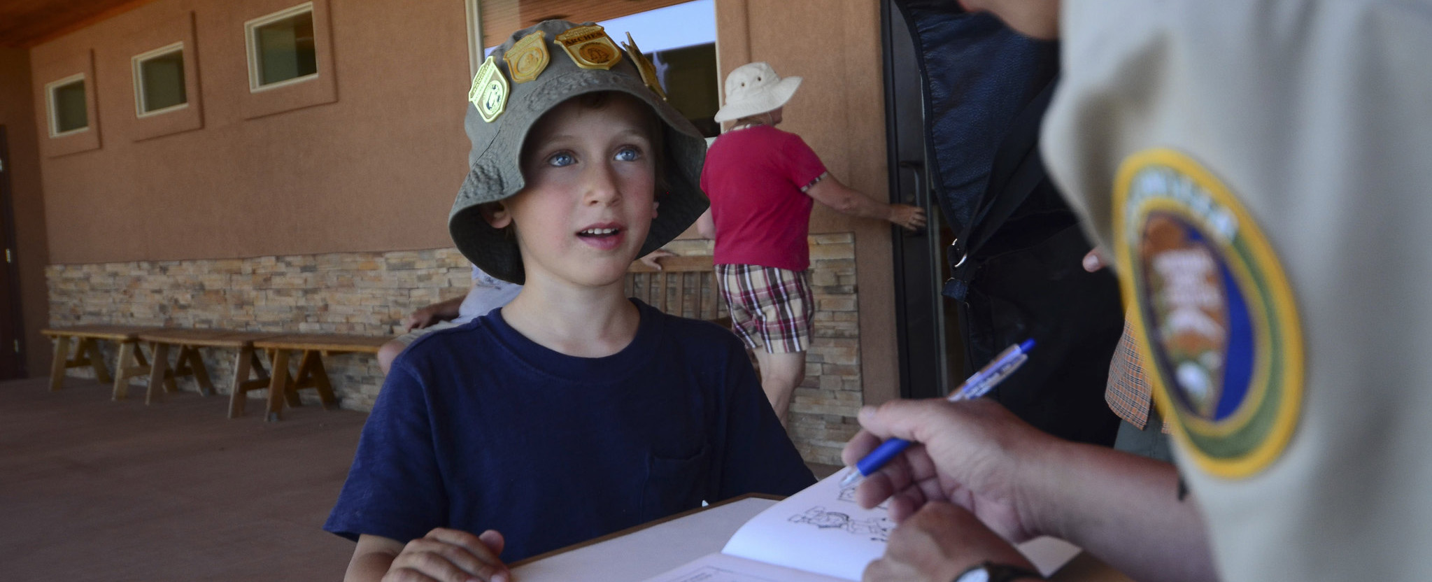 Junior Ranger wearing a hat with multiple Junior Ranger Badges reviews his book at the visitor center desk.