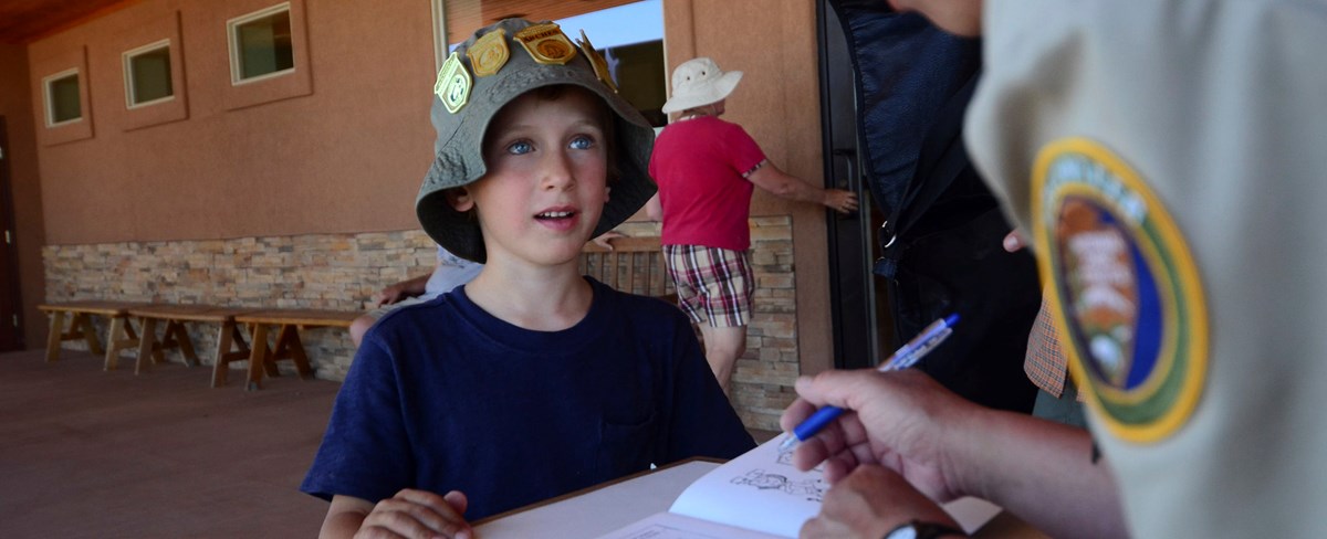 Junior Ranger wearing a hat with multiple Junior Ranger Badges reviews his book at the visitor center desk.