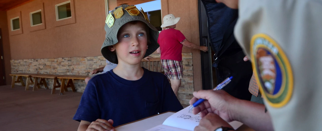 Junior Ranger Booklet Review Junior Ranger wearing a hat with multiple Junior Ranger Badges reviews his book at the visitor center desk.