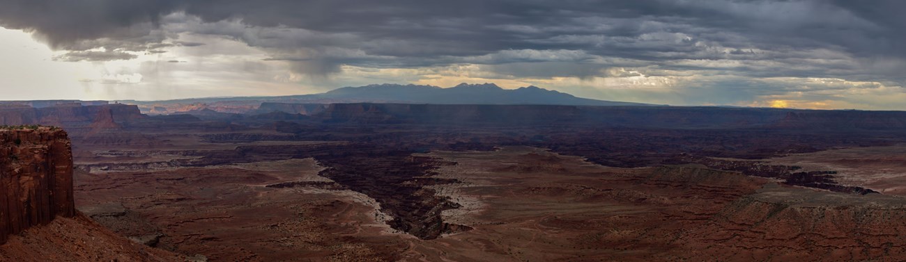 Dark blue storm clouds build above a sweeping overlook of mesa tops, mountains, and canyons.