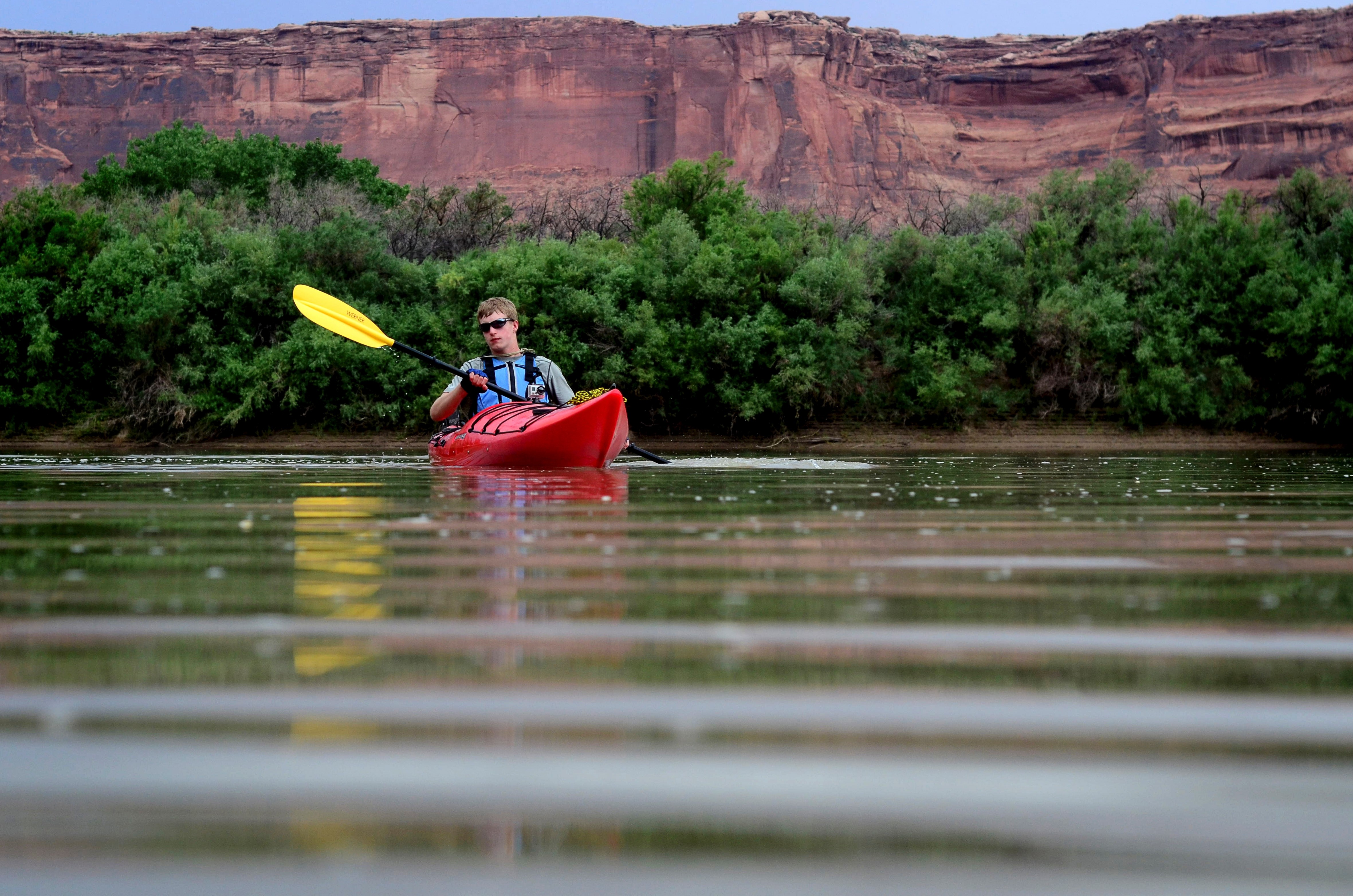 light-skinned man in red kayak paddles on a river, red canyon wall and green shrubs in background light-skinned man in red kayak paddles on a river, red canyon wall and green shrubs in background