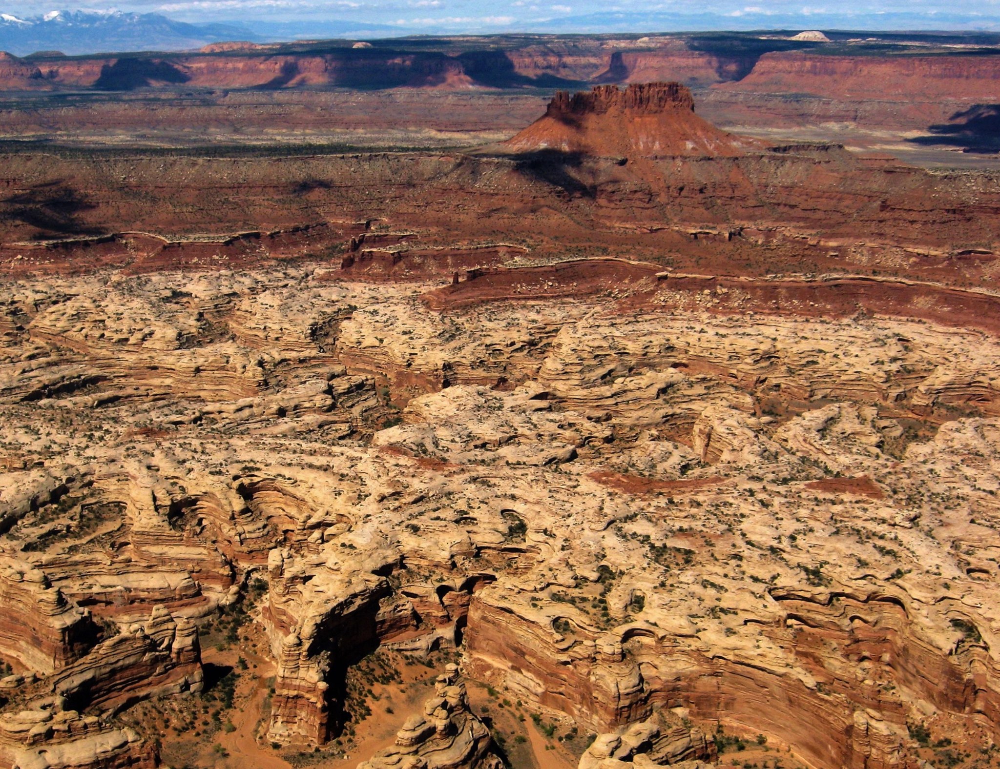 An aerial photo shows stripes and swirls of red and tan sandstone overlapping in a convoluted canyon. A large red butte stands tall in the background along the blue horizon.