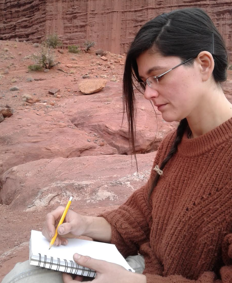 Jess Hough sketches using a pencil and notebook in front of reddish-orange cliffs.