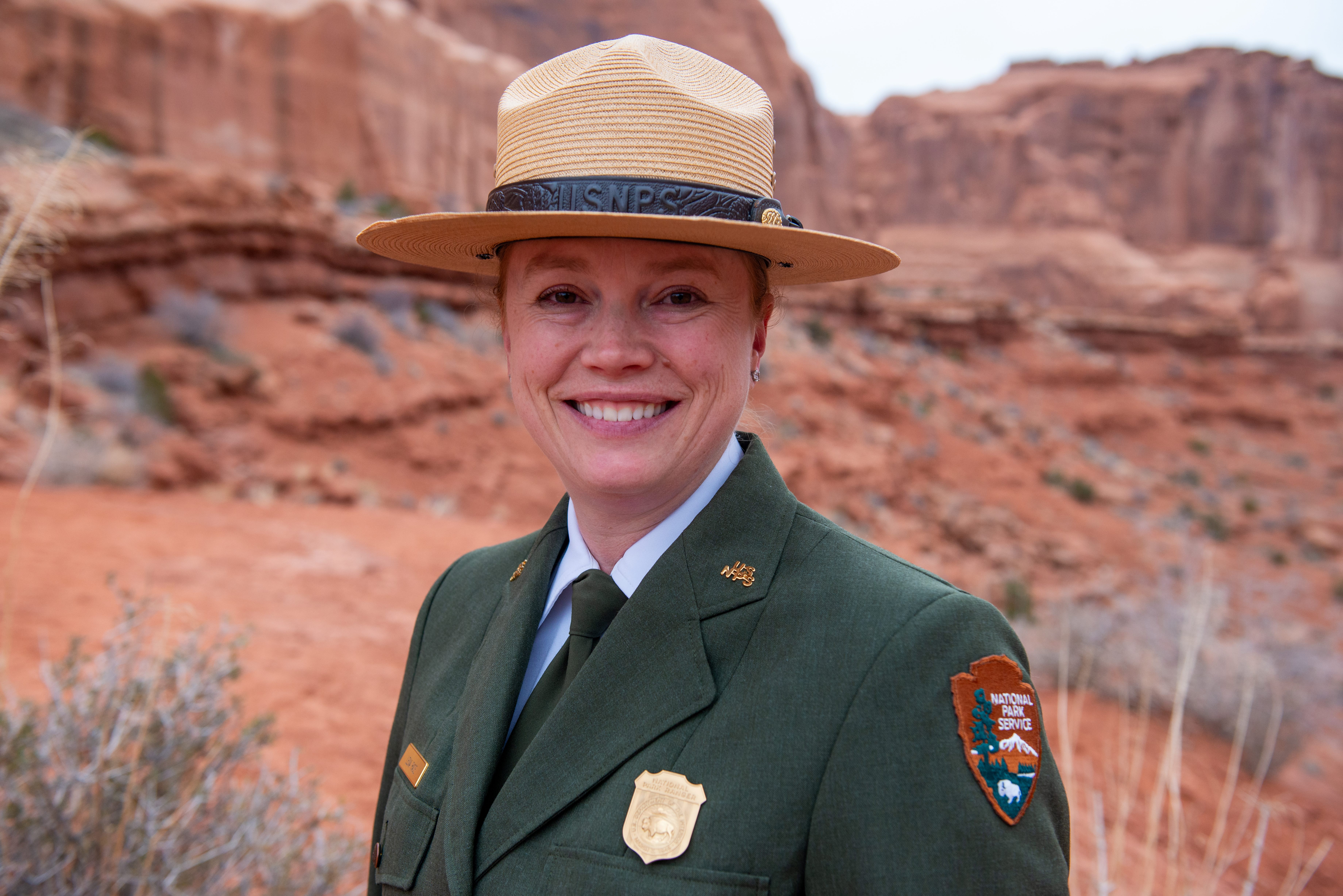 Smiling pale-skinned woman in green NPS dress uniform and NPS straw hat stands in front of red sandstone cliffs Smiling pale-skinned woman in green NPS dress uniform and NPS straw hat stands in front of red sandstone cliffs