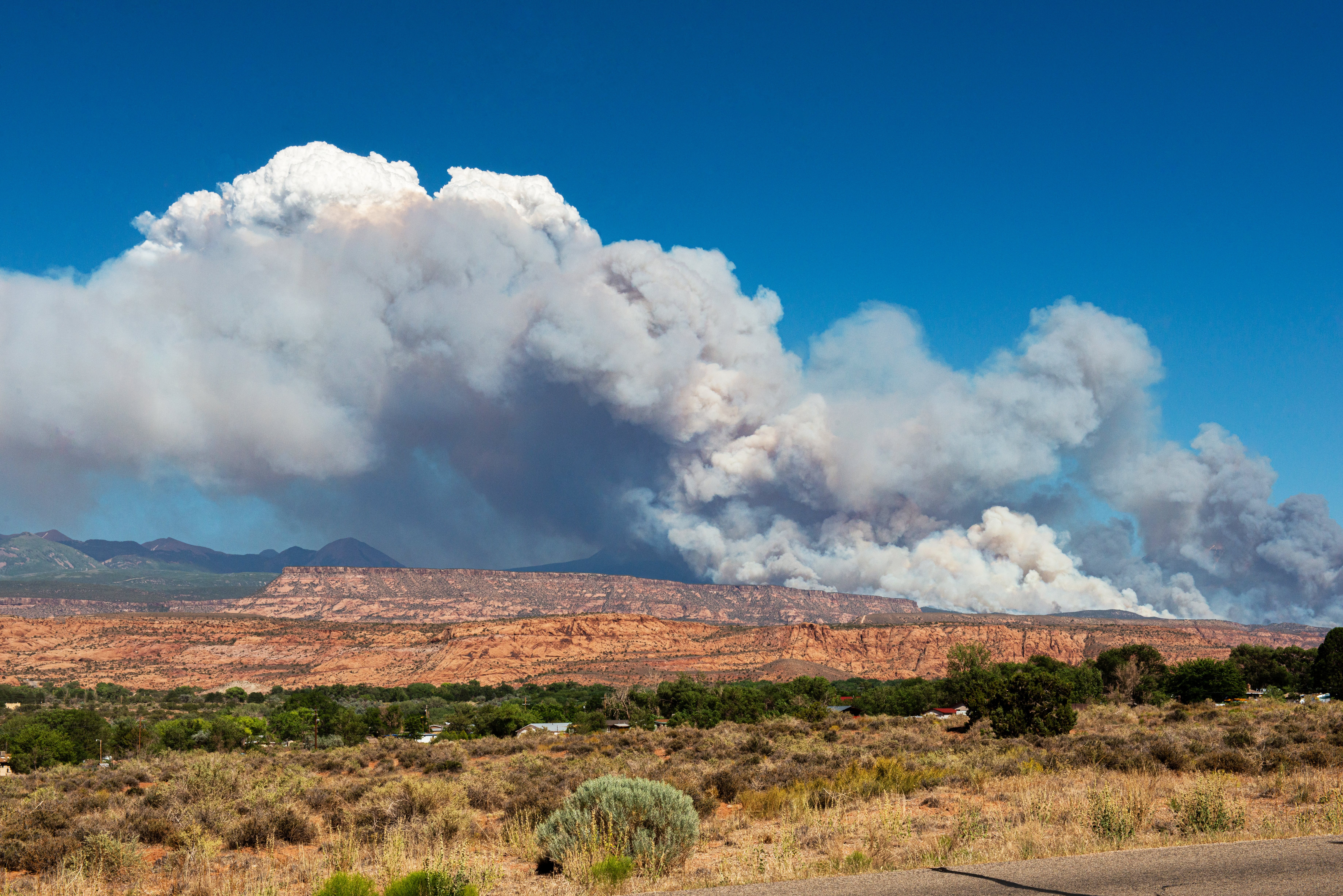 Dark gray and white clouds billow above a fire nestled in between mountains and orange and red sandstone ridges. In the foreground, shrubs and a few houses are visible.