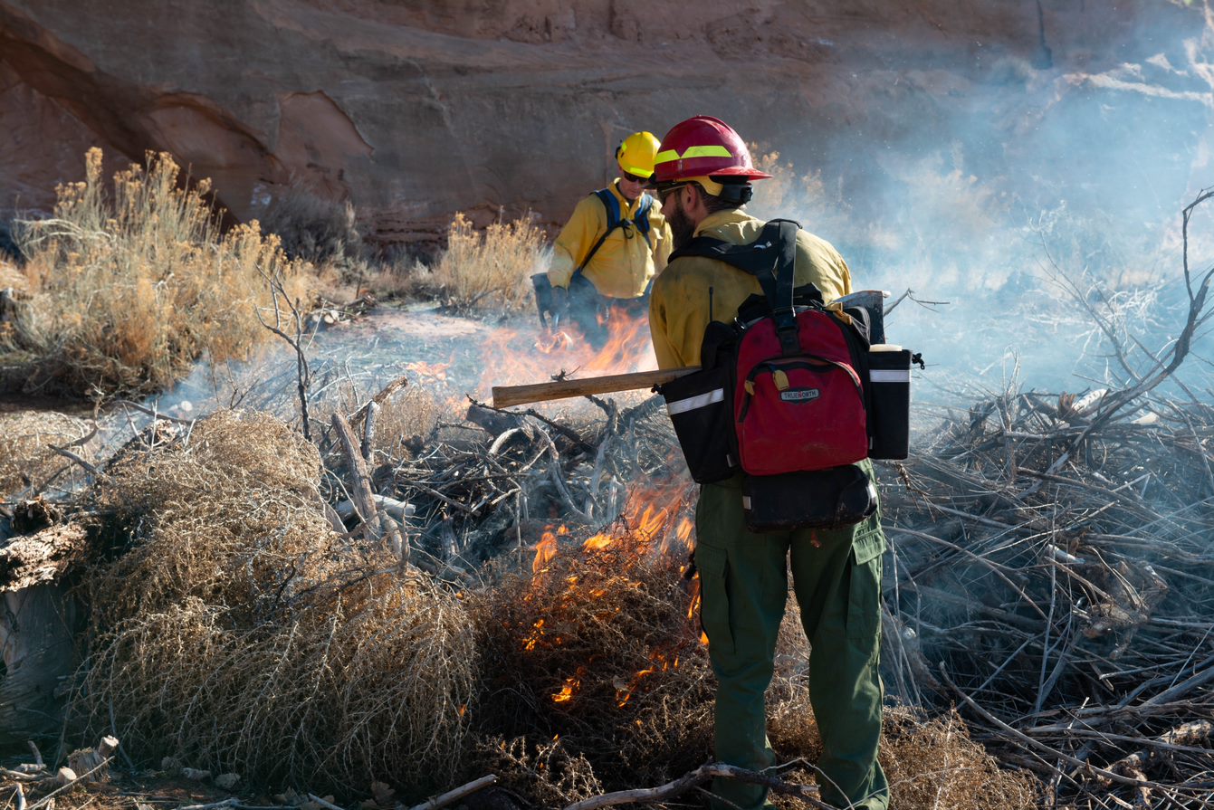 Prescribed burn in Arches National Park. NPS photo. Two male park rangers dressed in bright yellow fire jackets and red and yellow helmets supervise a large pile of burning vegetation.