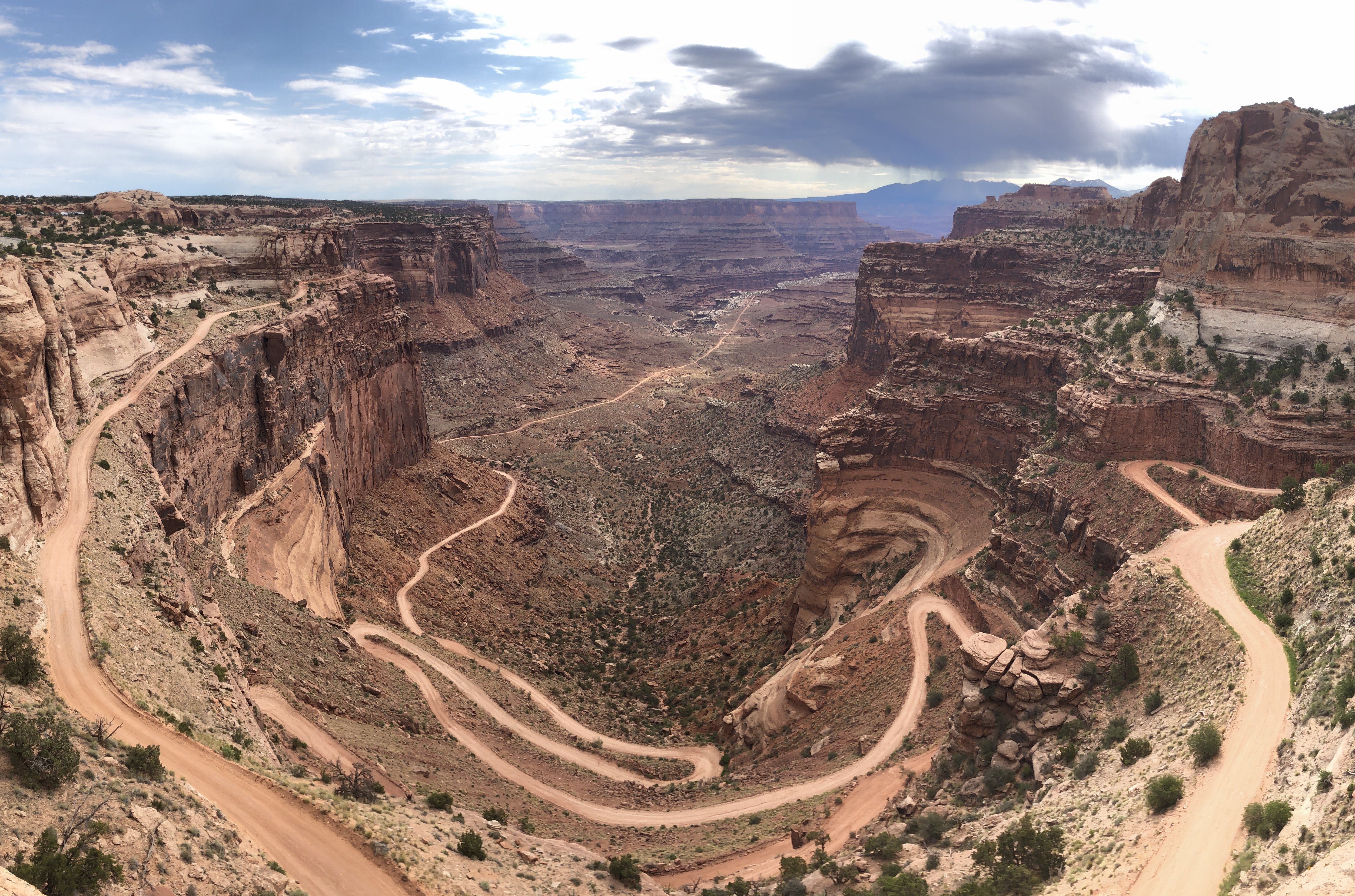 A winding reddish-orange road with switchbacks snakes its way down orange and brown canyon cliffs with a stormy mountain range in the background.