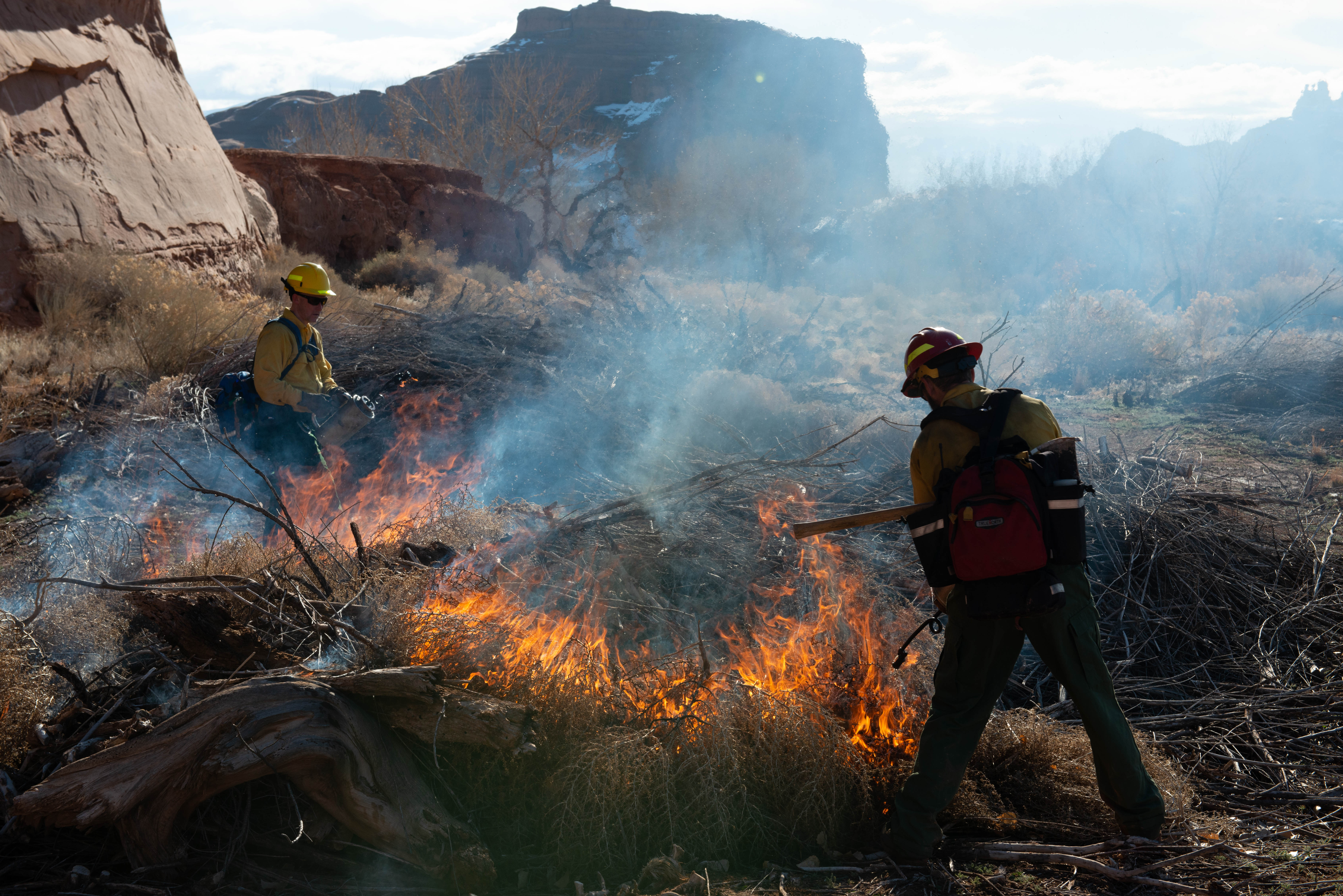 two people in yellow shirts and red hardhats use drip torches to ignite a pile of brush