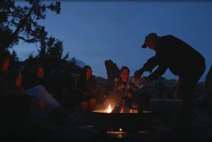 campers sit around a campfire in a fire ring while a standing person pours water onto it
