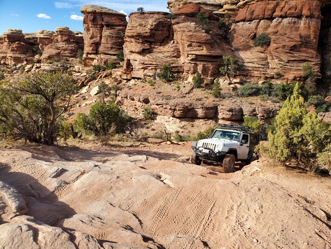 A white Jeep travels up a steep unpaved road with rock steps and outcroppings. Canyon walls surround the road.