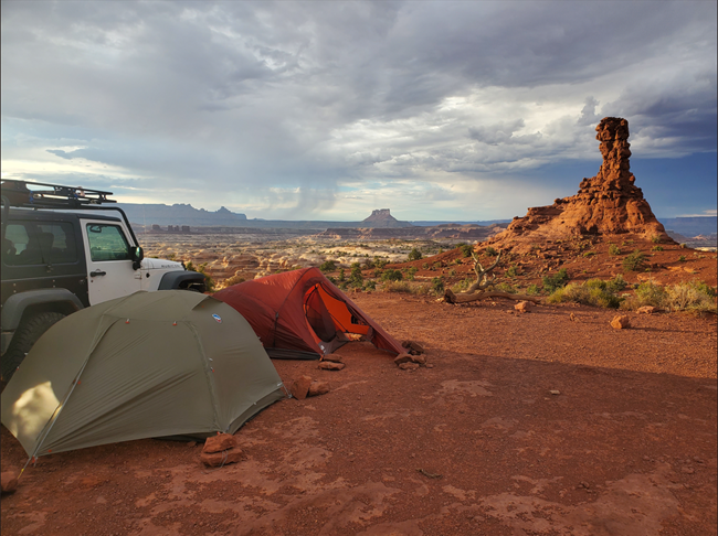 One green and one red tent are pitched next to a white Jeep. Sandstone formations surround the dirt campsite. Canyon walls and a rainstorm are in the distance.