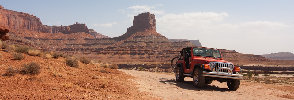 a red jeep drives a rock road in front of a tall rock tower