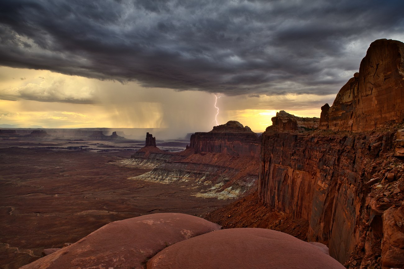 A monsoon storm over a desert landscape. Lightning strikes near tall sandstone formations. Canyons continue into the horizon.