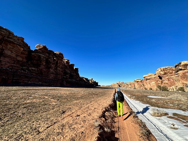 A backpacker walks along a dirt road through canyons with snow on the ground