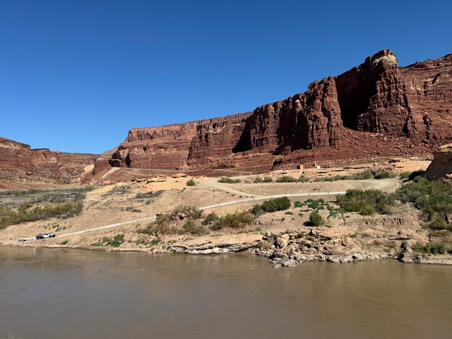 Boat launch with a steep ramp descending into the left corner of the image. Red rocks and sediment are in the foreground by a river bank. Cliff walls are in the background.