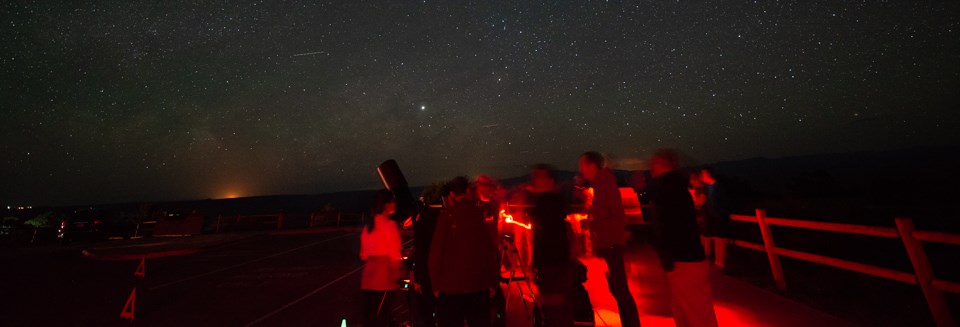 people illuminated with red lights stand near a telescope with a starry sky overhead