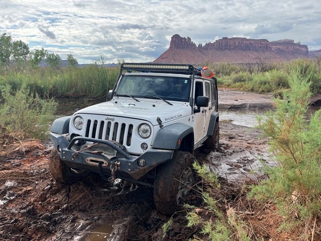Muddy conditions in a canyon wash with a creek crossing. A white Jeep is in the foreground with mud on the bottom.