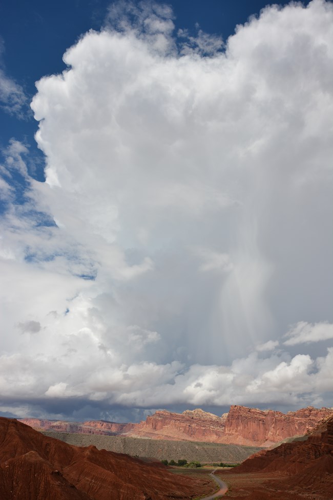 Towering white and gray clouds above colorful cliffs and a road.