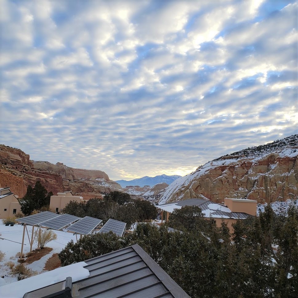 Adobe buildings with metal roofs and small windows, behind several solar panels, nestled below colorful red and white cliffs with a cloudy sky above.