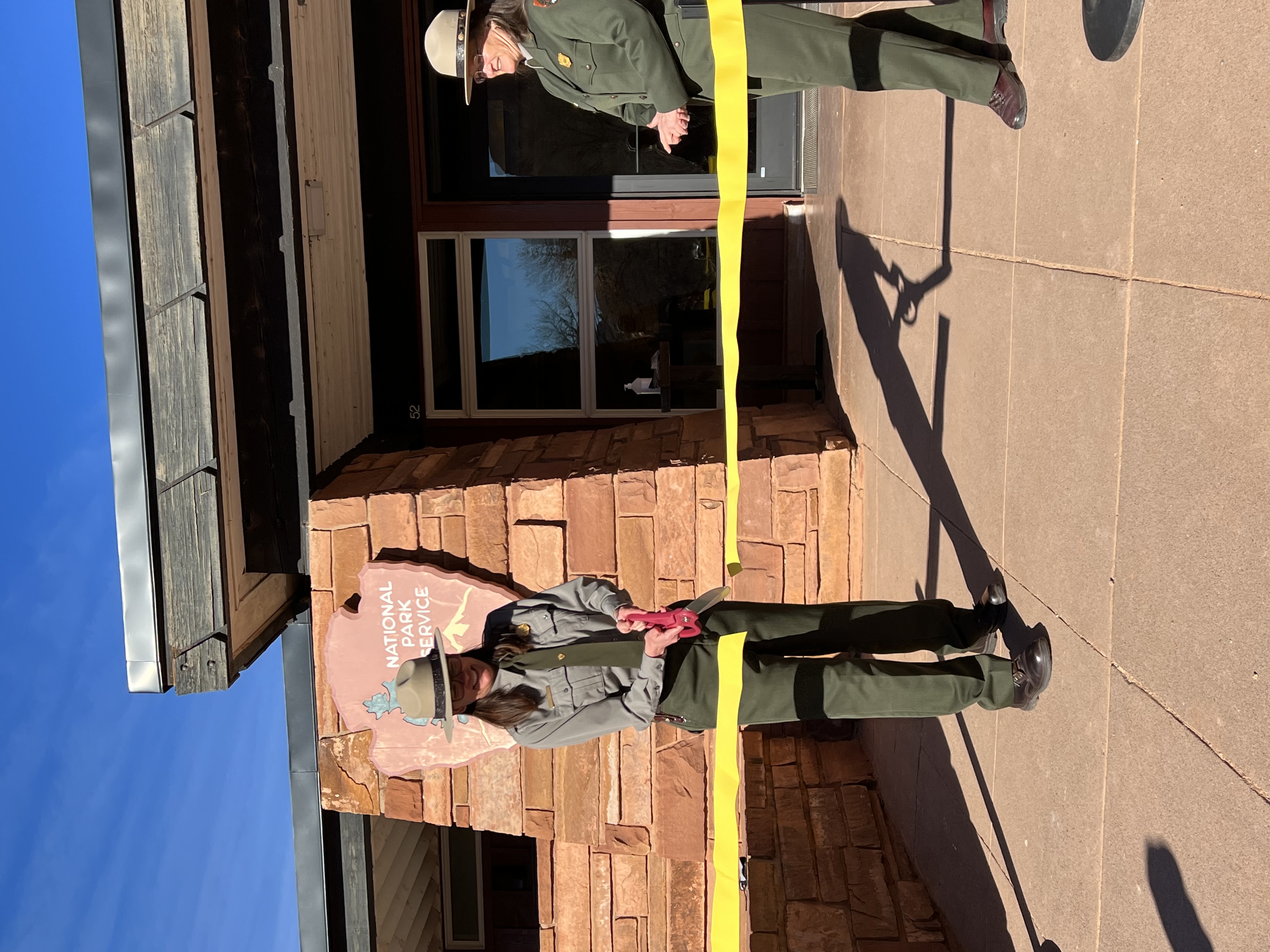 Two people stand in front of a building. One cuts a ribbon with oversized scissors