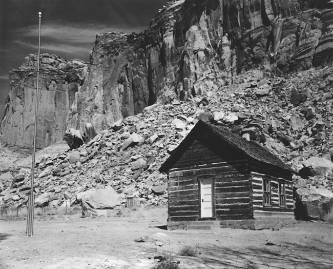 Black and white photo of log cabin building with a flagpole in front of it, and cliffs behind it.