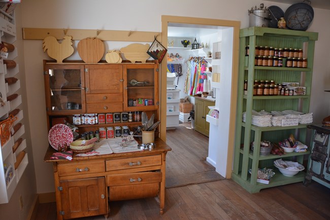 Room with wooden floors, a dresser, a bookshelf with sales items on them, looking into another room with aprons hanging on the walls.