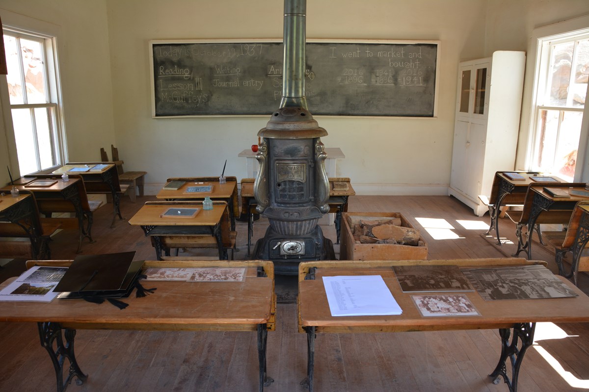 Old-fashioned desks, tables, and wood-burning stove inside a small building with with walls.